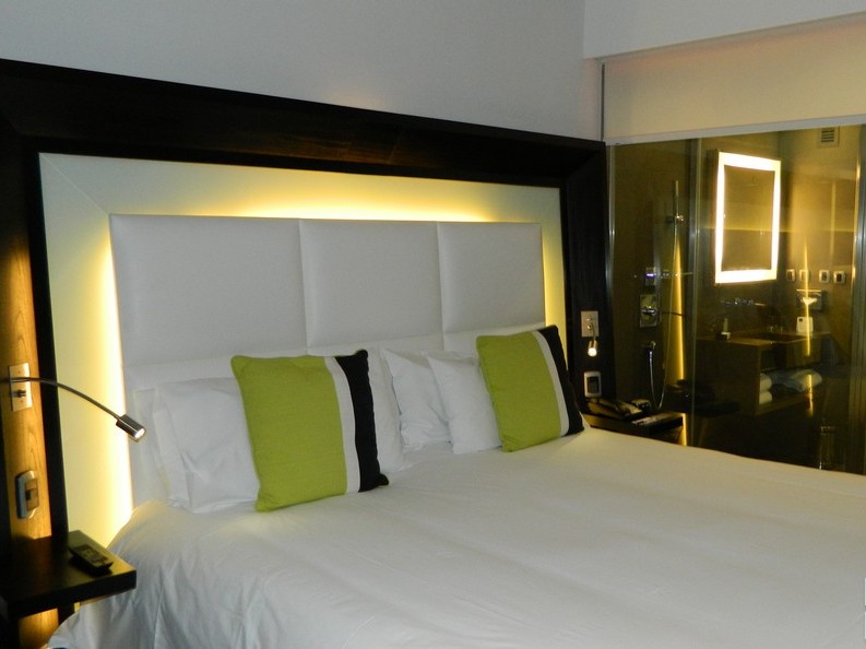 Uma das Suites Novotel, cama com iluminao e ao fundo box para banho de vidro 