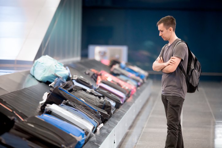 Young handsome man passenger in his 20s with carry-on backpack waiting at conveyor belt to pick his luggage in arrivals lounge of airport terminal building